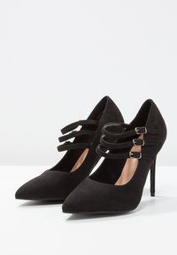 Svarta mocka högklackade skor med spetsig tå och tre dekorativa remmar, prydda med silverfärgad spännen och en hög stilettklack.
