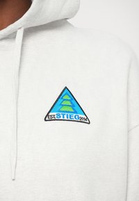 Gros plan sur un sweat à capuche blanc avec un logo triangulaire bleu et vert et le texte "EST. STIEG 2014" brodé sur la poitrine.