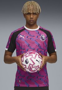 Tricou Puma cu model violet și roz, cu mâneci negre, având un logo cu leu. Ținând o minge de fotbal albă și roz cu design geometric.