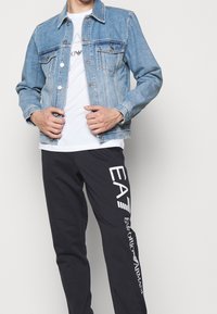 Světle modrá džínová bunda se dvěma náprsními kapsami; pod ní bílý tričko. Černé tepláky s bílým logem "EA7" po straně.