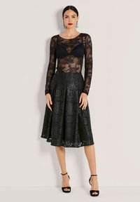 Zwart kanten langemouwen top over een zwarte bloemen midi rok met plooien. Accessoires zijn onder andere hangende oorbellen en zwarte gehakte sandalen.