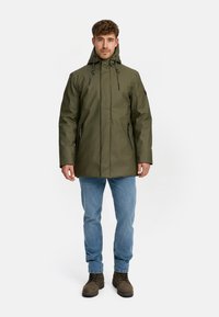 Khaki waterdichte parka met een capuchon, voorzien van twee met ritssluitingen afgesloten zakken, verstelbare toggles en een gladde textuur. Gecombineerd met lichtblauwe jeans en bruine laarzen.