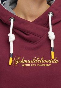Sudadera color burdeos con cordones blancos que presentan puntas amarillas y grises, y texto bordado en dorado que dice "Schmuddelwedda WENN DAT PLADDEERT."