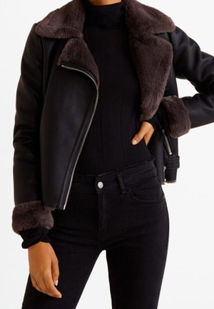 Person trägt eine schwarze Lammfelljacke mit dunkelbraunem Pelzkragen und Ärmelbündchen, kombiniert mit einem schwarzen Rollkragenpullover und schwarzen hochgeschnittenen Jeans.