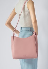Borsa a tracolla in pelle rosa con una texture liscia, bordo superiore curvo e tracolla regolabile. Presenta una tasca laterale e un marchio discreto alla base.