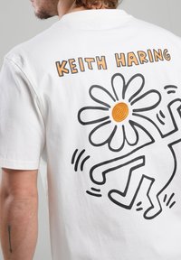 Camiseta blanca de algodón con un gráfico de contorno negro de una figura bailando y una gran margarita con un centro naranja. El texto dice "KEITH HARING".