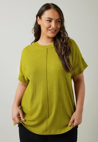 Top corto de manga corta en verde lima con una tela texturizada, que presenta un cuello redondo y un sutil detalle de costura central. Combinado con pantalones negros.
