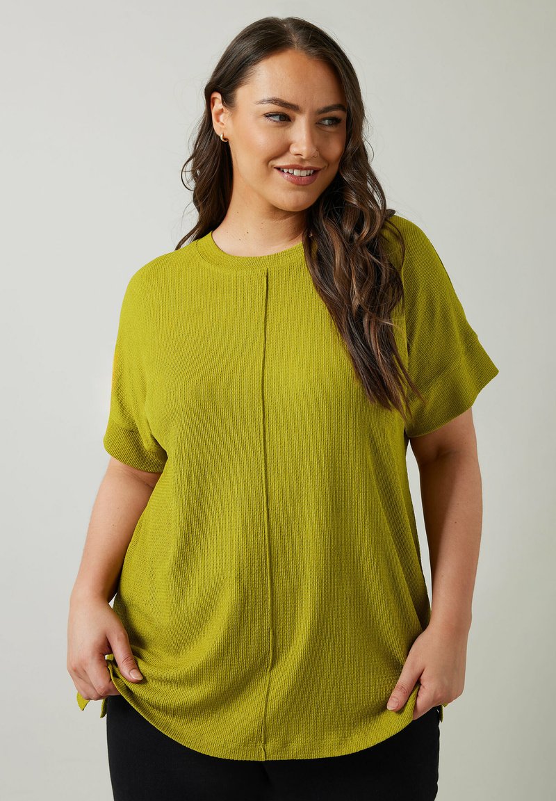 Top corto de manga corta en verde lima con una tela texturizada, que presenta un cuello redondo y un sutil detalle de costura central. Combinado con pantalones negros.
