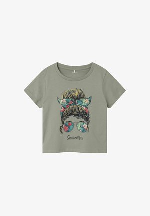 Zielona bawełniana koszulka z krótkim rękawem z grafiką przedstawiającą osobę w kwiecistej opasce na głowie i okularach przeciwsłonecznych. Poniżej znajduje się napis "Letnie Wibracje".