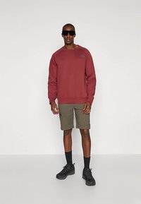 Sudadera burdeos con puños acanalados, pantalones cortos verde olivo con bolsillos laterales, calcetines negros y zapatos negros; modelo lleva gafas de sol.