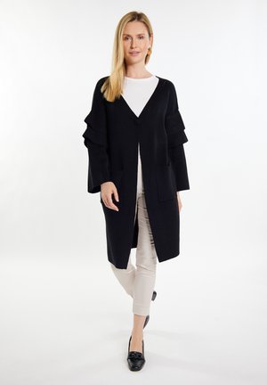 Langer schwarzer Cardigan mit V-Ausschnitt, Knopfverschluss und rüschigen Ärmeln. Getragen über einem weißen Oberteil, kombiniert mit hellen Hosen und schwarzen Schuhen.