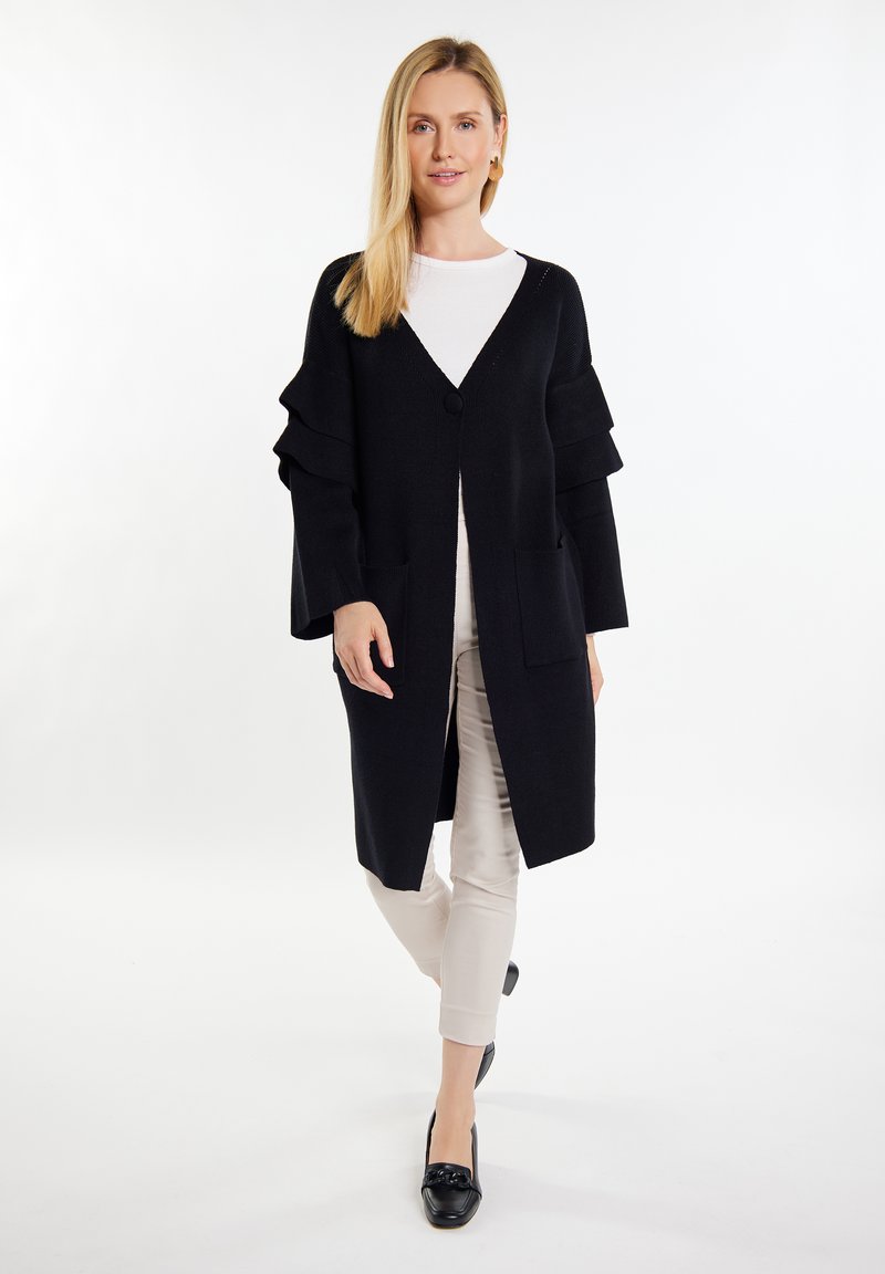 Cardigan lungo nero con scollo a V, chiusura a bottone e maniche arricciate. Indossato sopra una maglietta bianca e abbinato a pantaloni chiari e scarpe nere.