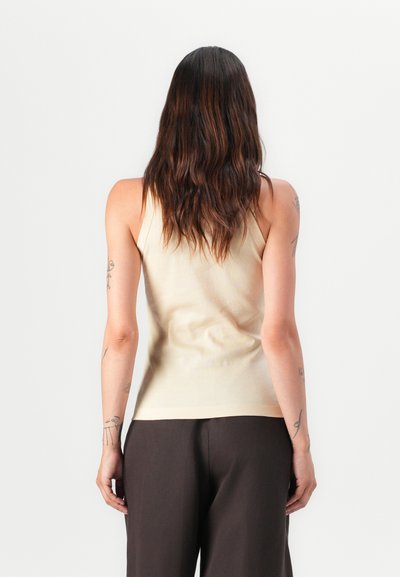 Top fără mâneci bej cu un design mulat, dotat cu bretele subțiri și o textură netedă, asociat cu pantaloni maro închis.