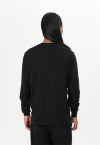 Zwarte lange mouwen T-shirt van katoen. Heeft een ronde hals en een kleurrafische opdruk op de rechter mouw. Glad textuur en een ontspannen pasvorm.
