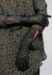 Černé kožené rukavice s hladkou texturou a přiléhavým střihem, nošené přes olivově zelenou blůzu s bílým abstraktním vzorem.