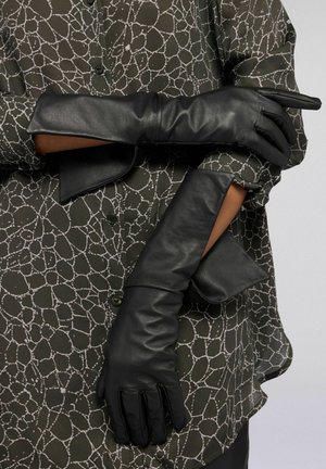 Czarne skórzane rękawiczki o gładkiej fakturze i dopasowanym kroju, noszone na oliwkowo-zielonej bluzce z białym abstrakcyjnym wzorem.
