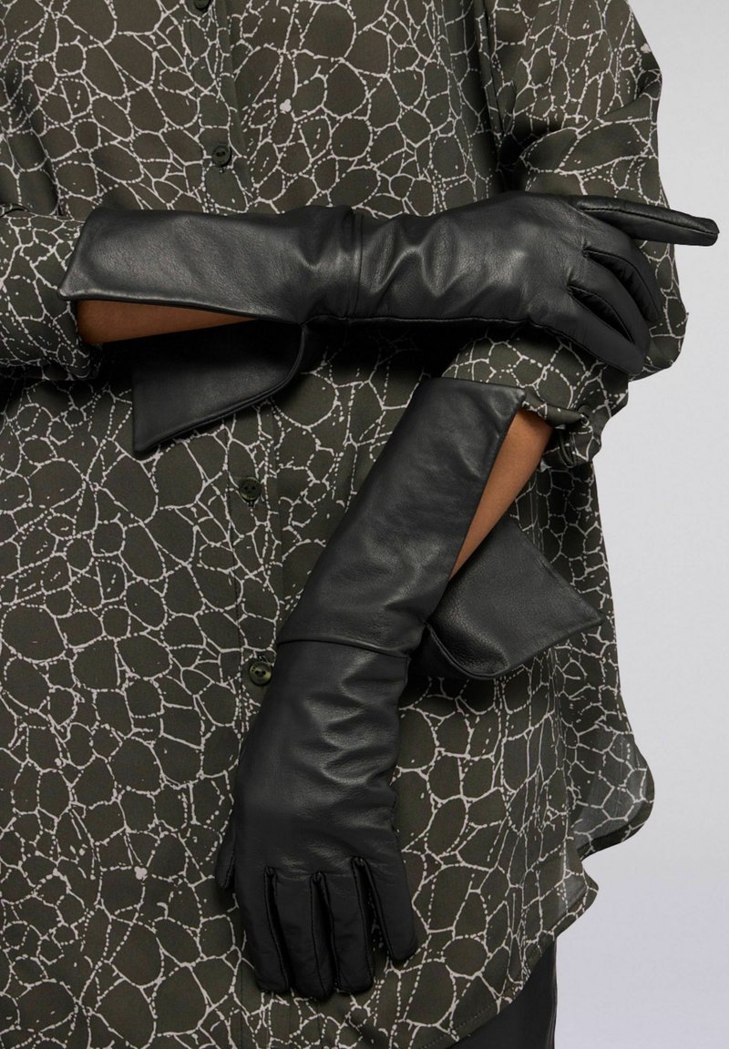 Černé kožené rukavice s hladkou texturou a přiléhavým střihem, nošené přes olivově zelenou blůzu s bílým abstraktním vzorem.