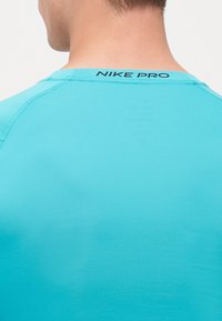 Turkis Nike Pro træningsskjorte, slim fit, glat tekstur, med en sort "NIKE PRO" logo ved baghalsen med flade sømme.