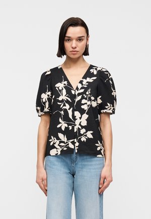 Žena v čiernej blúzke s gombíkmi s béžovým kvetinovým vzorom a nafúknutými krátkymi rukávmi, v kombinácii so svetlomodrými džínami, na jednoliatom pozadí.