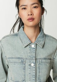 Giacca di jeans blu chiaro con colletto, chiusura anteriore con bottoni e due tasche sul petto. Presenta una texture sbiadita e cuciture a contrasto.