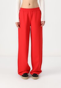 Pantalons à jambes larges en tissu rouge vif, dotés d'une taille élastique et d'une texture lisse. Associés à des chaussures noires et beige.