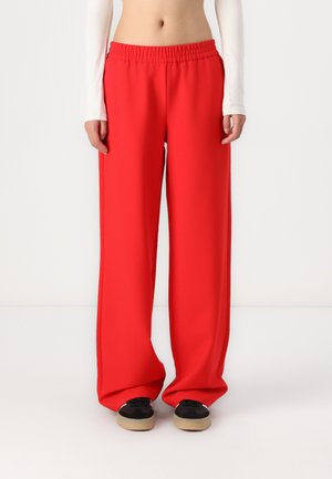 Pantalons à jambes larges en tissu rouge vif, dotés d'une taille élastique et d'une texture lisse. Associés à des chaussures noires et beige.
