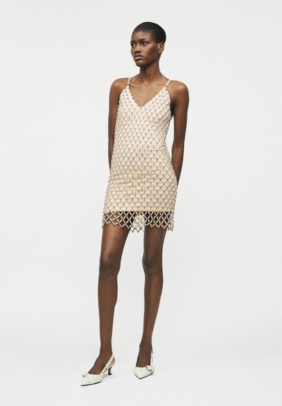 Γυναίκα που φοράει μια μπεζ φόρεμα slip καλυμμένη με δίχτυ από χάντρες, συνδυασμένη με λευκά slingback παπούτσια με μικρό τακούνι kitten heel, στέκεται μπροστά σε απλό φόντο.