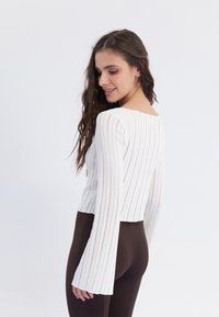 Fehér csíkos kötött crop top hosszú, harang alakú ujjakkal. A kialakításon széles nyakkivágás és textúrált anyag található. Barna leggings-szel párosítva.