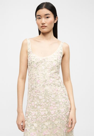 Νέα γυναίκα με ίσια μαύρα μαλλιά, φορώντας αμάνικο μπεζ φόρεμα με ροζ και λευκή κεντητική ανθοδέσμη, που στέκεται μπροστά σε απλό φόντο.