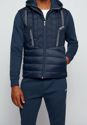 Homme portant une veste matelassée à capuche bleu marine par-dessus un haut à manches longues assorti et un pantalon de jogging avec le logo BOSS sur la poitrine et la cuisse.