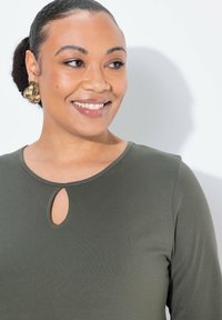 Olivgrön långärmad topp med rund halsringning och nyckelhålsdetalj. Modellen bär ett stort, texturerat guldörhänge. Slät tygfinish.