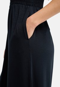 Χέρι που μπαίνει στην πλαϊνή τσέπη μιας μαύρης φούστας με ελαστική μέση, εμφανίζοντας την υφή του υφάσματος και τη λεπτομέρεια του ανοίγματος της τσέπης.