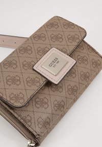 Sac bandoulière marron Guess avec motif logo répété, fermeture à rabat et plaque métallique indiquant "Guess EST 1981" à l’avant.