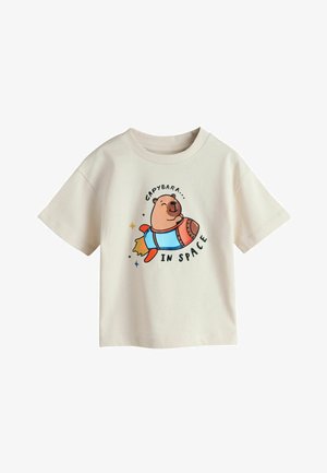 Krémová košeľa s krátkym rukávom so usmievajúcou sa kapibarou na rakete, s textom „Kapibara vo vesmíre“ okolo ilustrácie.