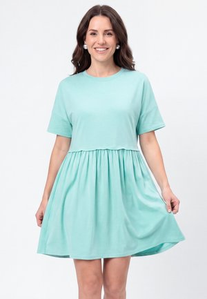 Mujer con cabello largo y oscuro sonriendo, lleva grandes pendientes de perlas y un vestido suelto de manga corta azul claro con falda recogida.