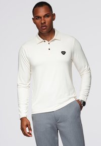 Polo a maniche lunghe di colore crema con colletto, dotato di tre bottoni scuri e un logo triangolare nero sul lato sinistro del petto. Indossato con pantaloni grigi.