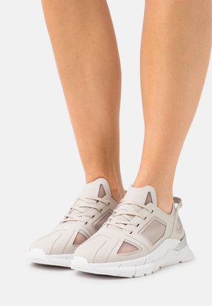Personne portant des baskets sportives beige clair avec des semelles blanches, se tenant sur un fond blanc uni.