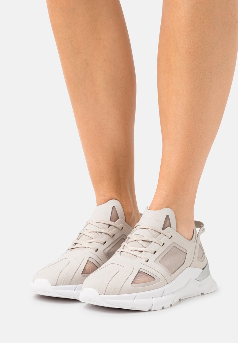 Person som bär ljusbeige sportskor med vita sulor, stående mot en helt vit bakgrund.