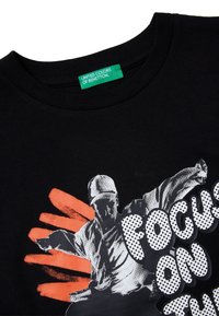 Černé bavlněné tričko s krátkými rukávy, s grafikou postavy a nápisem "FOCUS ON" tučným, puntíkatým písmem na oranžovém pozadí.