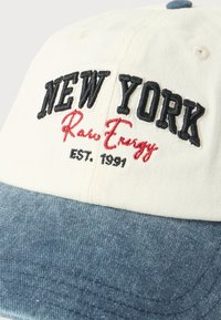 Λευκό και μπλε καπέλο μπέιζμπολ με κεντημένη τη φράση "NEW YORK Raw Energy EST. 1991" με μαύρη και κόκκινη κλωστή στον μπροστινό πίνακα.