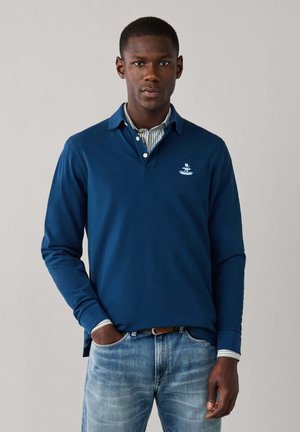Hombre con un polo azul de manga larga sobre una camisa de rayas, con vaqueros azul claro y una mano en el bolsillo, de pie frente a un fondo liso.