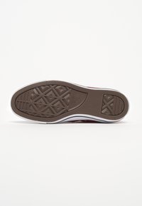 Semelle en caoutchouc marron d'une chaussure Converse All Star avec des crampons à motifs et une semelle intermédiaire blanche visible sur un fond blanc uni.