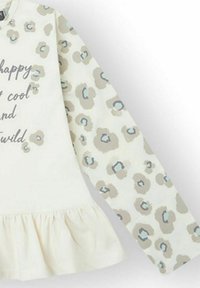 Camiseta de manga larga en color crema con manchas de leopardo marrón claro, con un dobladillo con volantes y texto impreso: "Feliz, genial y salvaje". Hecha de tela suave.