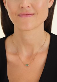 Collana in oro con pendente a forma rettangolare; superficie liscia, catena sottile con piccoli anelli rotondi, indossata sopra un top scuro.