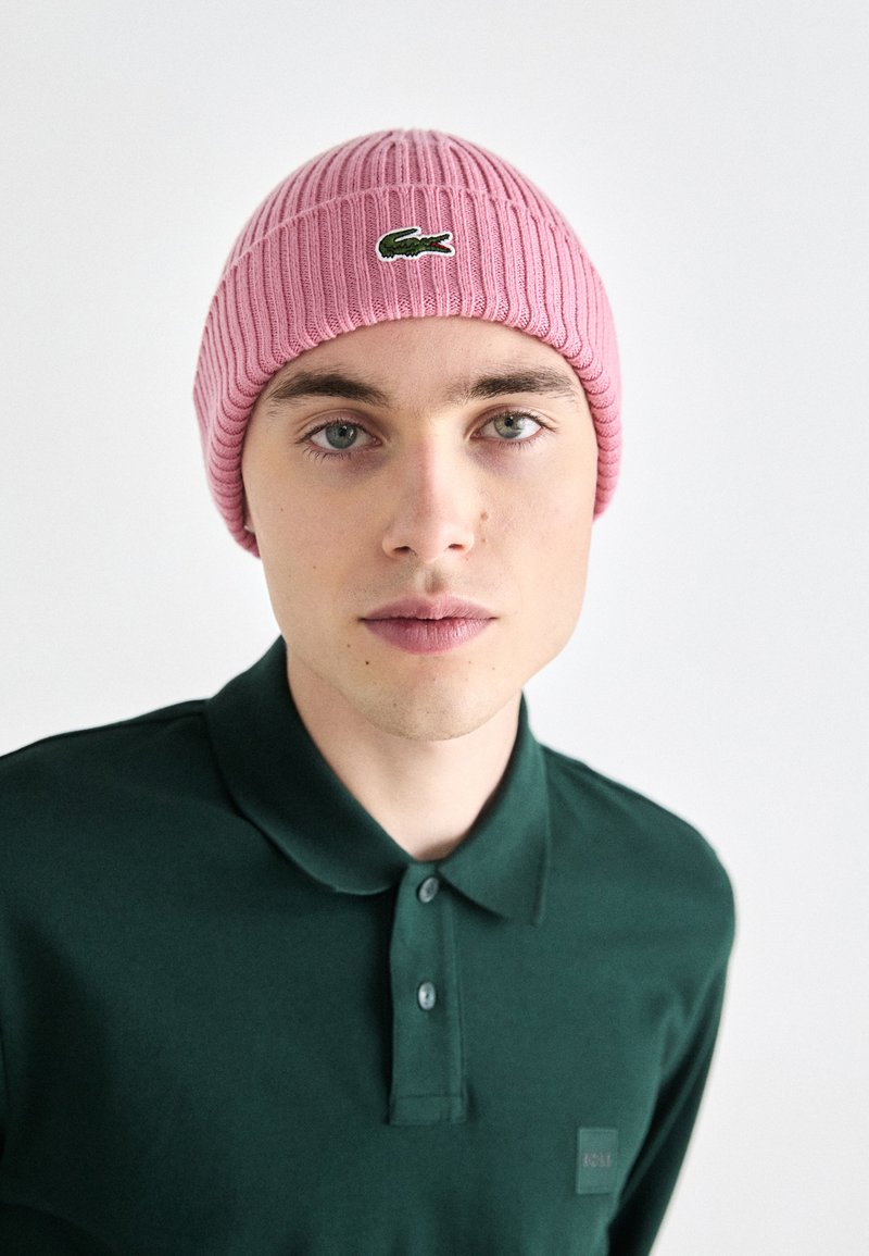 Różowa czapka dziana z prążkowanym wzorem i zielonym logo, noszona przez osobę w ciemnozielonej koszulce polo. Prosty, codzienny design.