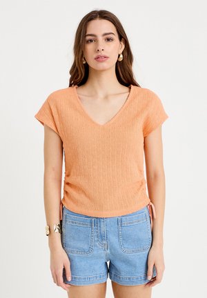 Kobieta ubrana w brzoskwiniowy, teksturowany top z krótkim rękawem i dekoltem w serek oraz jasnoniebieskie szorty jeansowe z dużymi przednimi kieszeniami, stojąca na białym tle.