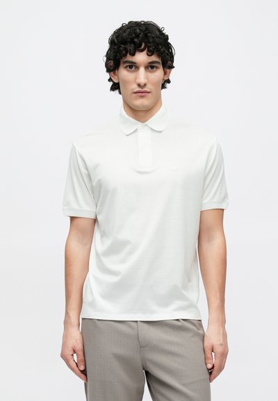 Bílá polokošile s plochým límcem, krátkými rukávy a jemným logem na hrudi. Materiál působí lehce a hladce. Nositelná s světle šedými kalhotami.