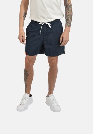 Uomo che indossa pantaloncini blu navy con coulisse, sneakers bianche e t-shirt bianca, mostrando tatuaggi su braccia e gamba destra.