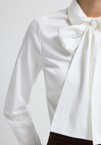 Witte blouse met een grote strikkragen, met lange mouwen en een gladde textuur, gecombineerd met een bruine gestructureerde onderkant.