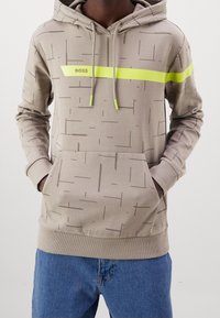 Beige hoodie med ett geometriskt mönster, med en ljusgul horisontell rand över bröstet och matchande gula dragsnören.